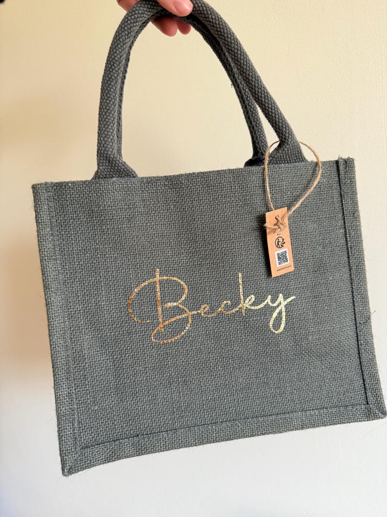 May include: A gray jute tote bag with a gold script name "Becky" on the front. The bag has matching gray handles and a small tag attached. The bag is held up by a hand.