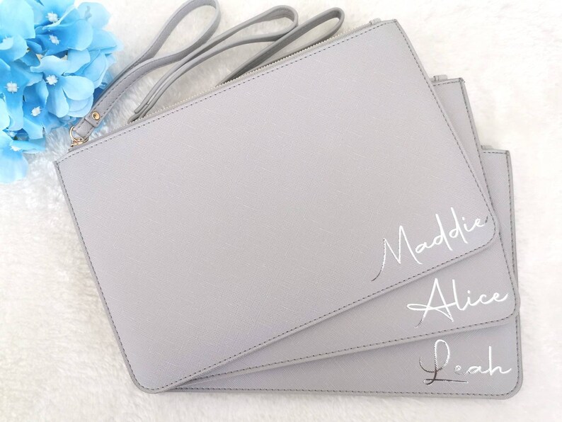 May include: Three gray zippered pouches with silver lettering. The pouches are stacked on top of each other. The top pouch has the name Maddie written on it. The middle pouch has the name Alice written on it. The bottom pouch has the name Leah written on it.