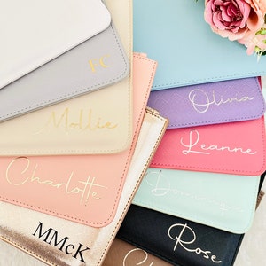 May include: A collection of personalized clutches in various colors, including white, gray, beige, pink, light blue, purple, red, mint green, black, and brown. Each clutch features a gold or black inscription with a name, such as "Mollie", "Olivia", "Leanne", "Charlotte", "Rose", and "Cherie".