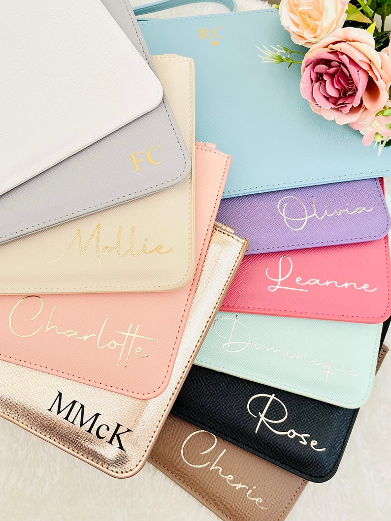 May include: A collection of personalized clutches in various colors, including white, gray, beige, pink, light blue, purple, red, mint green, black, and brown. Each clutch features a gold or black inscription with a name, such as "Mollie", "Olivia", "Leanne", "Charlotte", "Rose", and "Cherie".