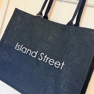 May include: A navy blue tote bag with the words "Island Street" in white lettering. The bag has two navy blue handles and is hanging on a white door with silver hooks.