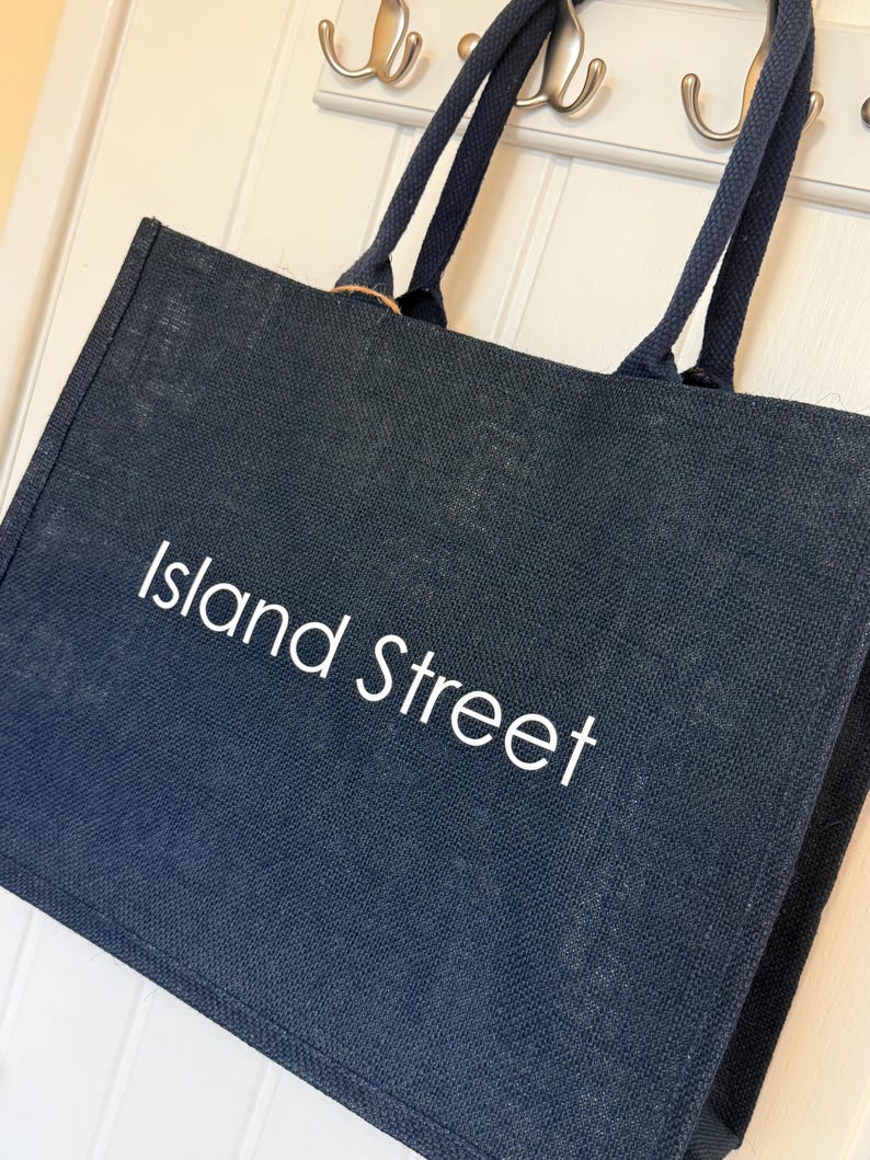 May include: A navy blue tote bag with the words "Island Street" in white lettering. The bag has two navy blue handles and is hanging on a white door with silver hooks.