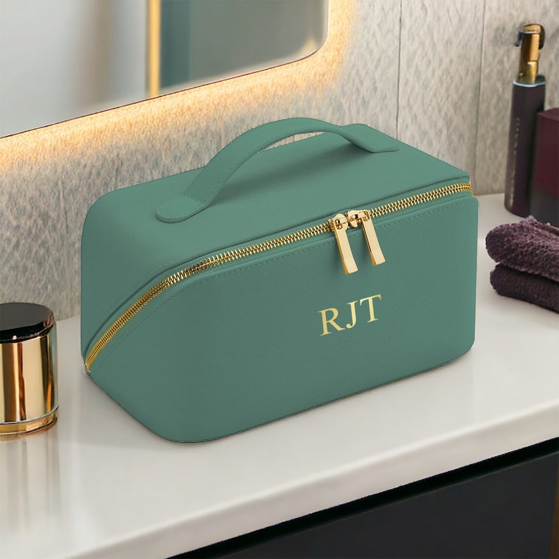 May include: Teal makeup bag with a gold zipper and handle. The bag has the initials "RJT" in gold lettering. The bag is sitting on a white surface next to a mirror and other bathroom accessories.