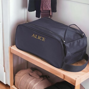 May include: A dark navy blue rectangular travel bag featuring the name ALICE embroidered in gold thread on the front side. The bag includes a top zip closure, a side pocket, and a sturdy fabric handle attached to the side for easy carrying.