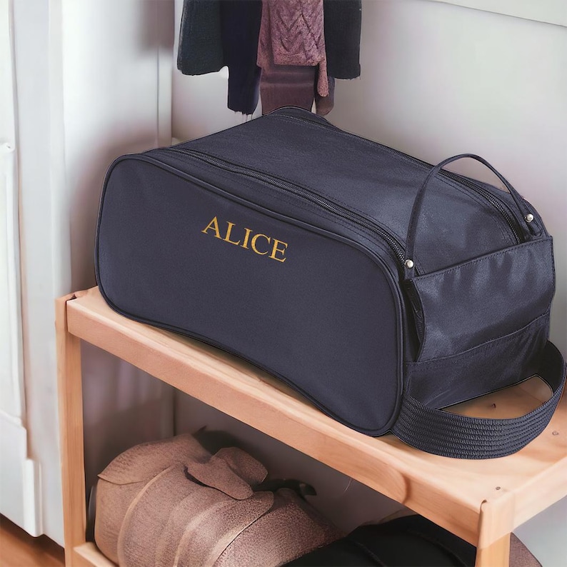 May include: A dark navy blue rectangular travel bag featuring the name ALICE embroidered in gold thread on the front side. The bag includes a top zip closure, a side pocket, and a sturdy fabric handle attached to the side for easy carrying.
