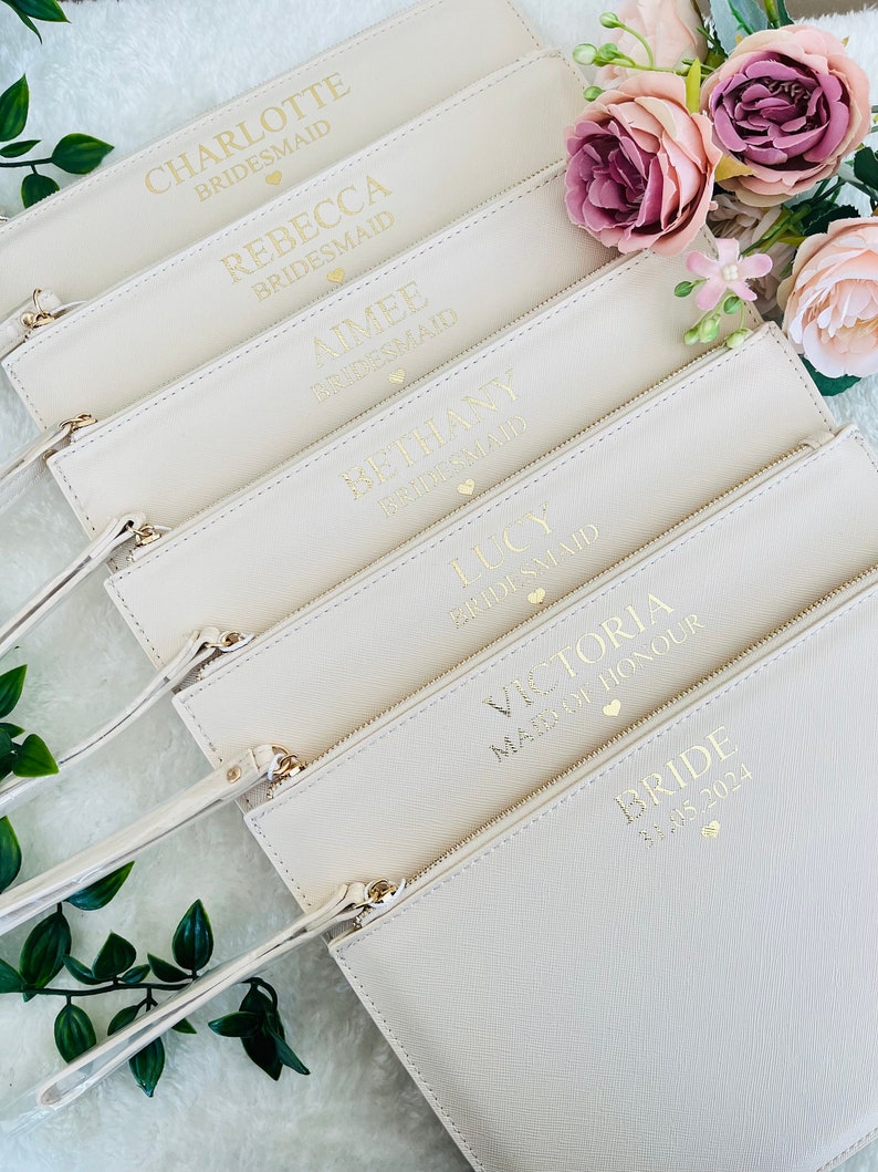 May include: A collection of cream-colored rectangular wristlet pouches with gold-toned zippers and wrist straps. Each pouch features gold foil text including names like Charlotte, Rebecca, Aimee, Bethany, Lucy, Victoria, and Bride, with small heart icons.
