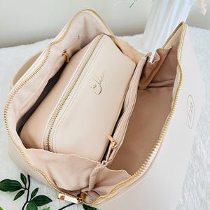 May include: Open cream-colored cosmetic bag with gold zippers and a matching smaller case inside. The smaller case has the name "Ellen" in gold script. The bag has a gold "EB" monogram on the side.