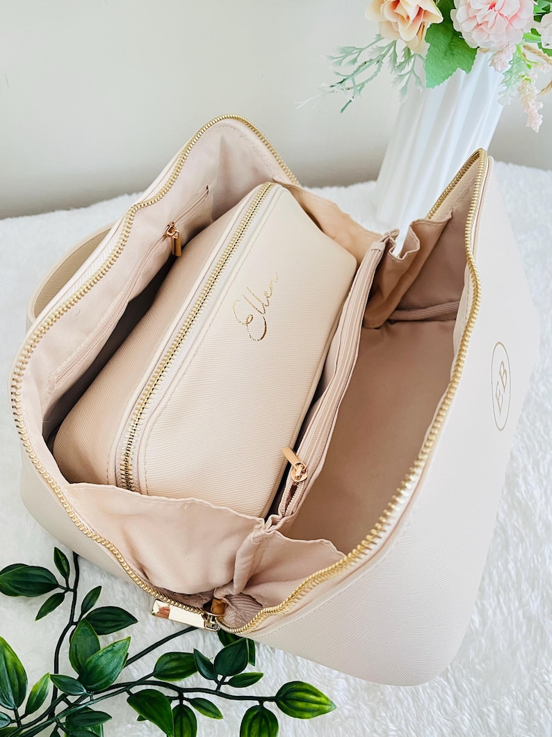May include: Open cream-colored cosmetic bag with gold zippers and a matching smaller case inside. The smaller case has the name "Ellen" in gold script. The bag has a gold "EB" monogram on the side.