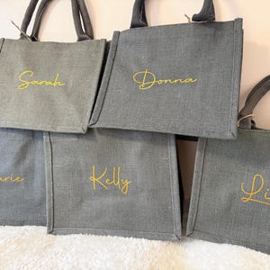 May include: A collection of gray tote bags with matching handles. Each bag has a name embroidered in gold script: Sarah, Donna, Marie, Kelly, and Lisa. The bags are made of a textured fabric and are likely for carrying items.