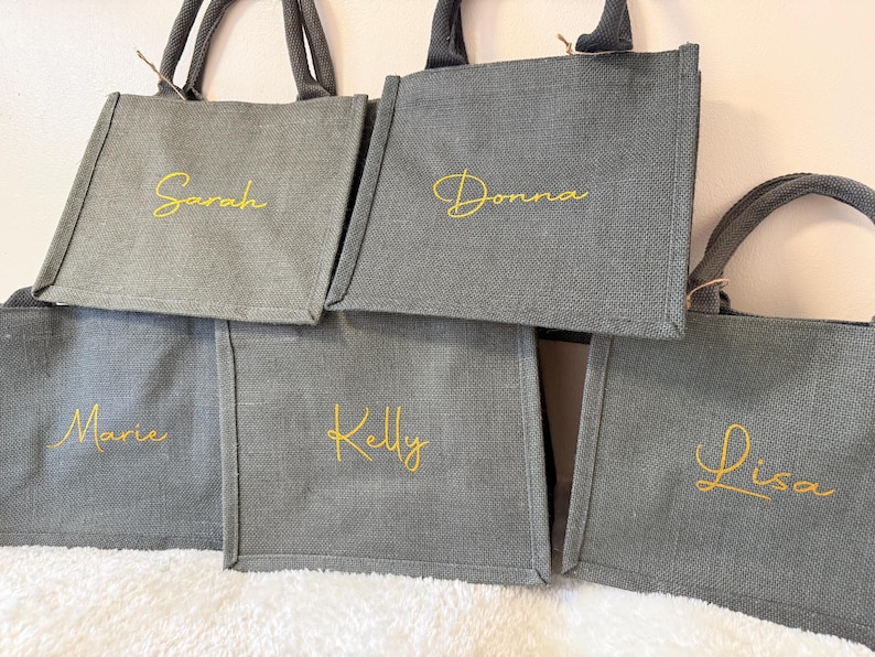May include: A collection of gray tote bags with matching handles. Each bag has a name embroidered in gold script: Sarah, Donna, Marie, Kelly, and Lisa. The bags are made of a textured fabric and are likely for carrying items.