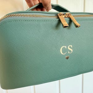 Pu&ograve; includere: Un beauty case verde acqua con cerniera e manico dorati. La borsa presenta le iniziali "CS" e un simbolo a cuore in oro. La borsa &egrave; tenuta in mano.