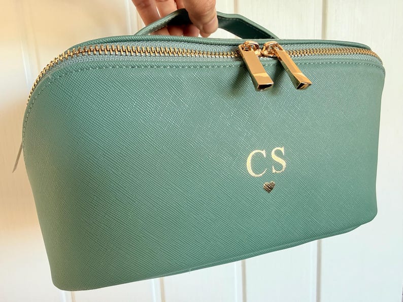 Pu&ograve; includere: Un beauty case verde acqua con cerniera e manico dorati. La borsa presenta le iniziali "CS" e un simbolo a cuore in oro. La borsa &egrave; tenuta in mano.