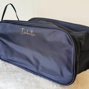 May include: A rectangular navy blue toiletry bag made of a smooth, durable fabric. The front panel features the name Isabella embroidered in a thin, metallic gold script font. The bag includes a side handle and a zip closure along the top edge.