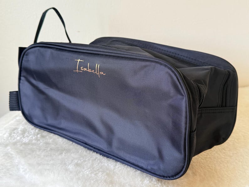 May include: A rectangular navy blue toiletry bag made of a smooth, durable fabric. The front panel features the name Isabella embroidered in a thin, metallic gold script font. The bag includes a side handle and a zip closure along the top edge.