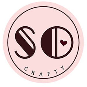 May include: A pink circle with the word "SO CRAFTY" in black text at the bottom. The letters "SO" are in a stylized font with black and white stripes. The letter "O" has a small red heart in the center.