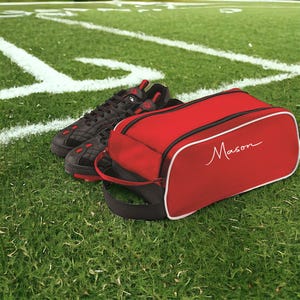 May include: A pair of black athletic shoes with red accents positioned next to a rectangular red zippered bag with white piping and the name Mason printed in white script on the side. The bag features a black handle on the side for easy carrying.