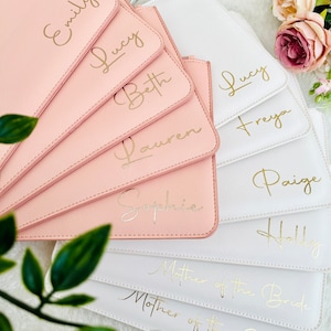 May include: A collection of rectangular pouches in blush pink and white, each featuring gold foil script lettering. The pink pouches are personalized with names Emily, Lucy, Beth, Lauren, and Sophie. The white pouches feature names Lucy, Freya, Paige, Holly, and the titles Mother of the Bride and Mother of the Groom.