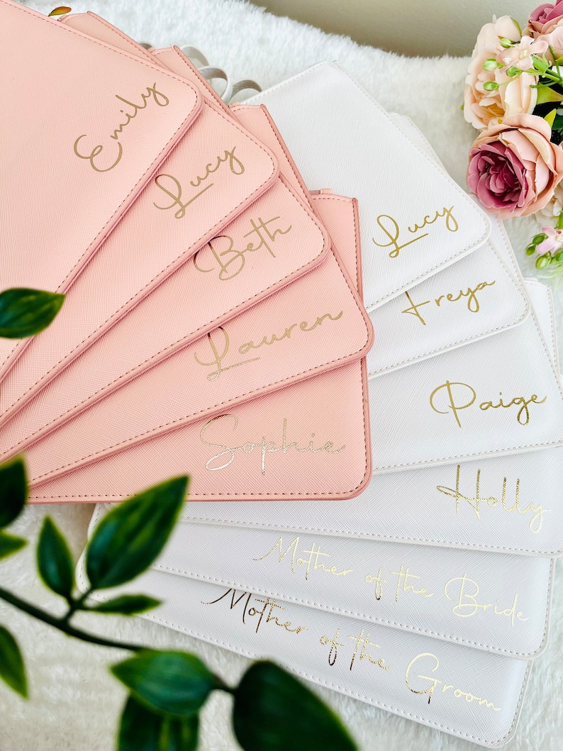 May include: A collection of rectangular pouches in blush pink and white, each featuring gold foil script lettering. The pink pouches are personalized with names Emily, Lucy, Beth, Lauren, and Sophie. The white pouches feature names Lucy, Freya, Paige, Holly, and the titles Mother of the Bride and Mother of the Groom.