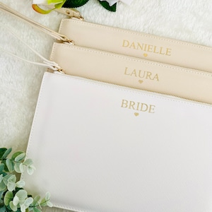 May include: Three white zippered pouches with gold lettering. The pouches are stacked on top of each other. The top pouch says "BRIDE" with a small heart, the middle pouch says "LAURA" with a small heart, and the bottom pouch says "DANIELLE" with a small heart.