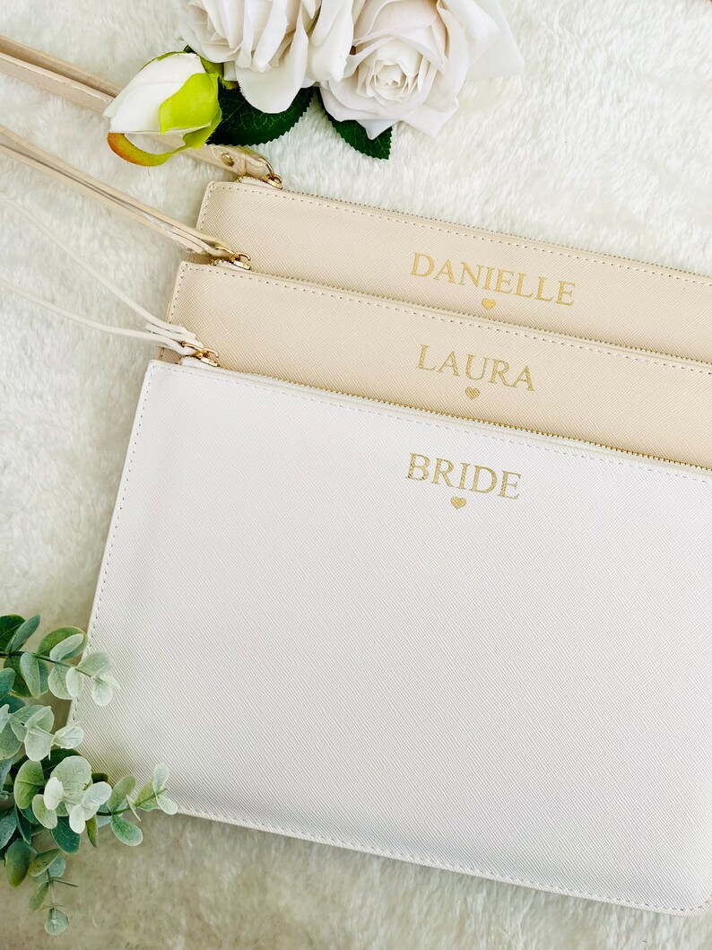May include: Three white zippered pouches with gold lettering. The pouches are stacked on top of each other. The top pouch says "BRIDE" with a small heart, the middle pouch says "LAURA" with a small heart, and the bottom pouch says "DANIELLE" with a small heart.