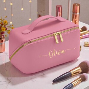 May include: A pink makeup bag with a gold zipper and handle. The bag has the name "Olivia" in gold script. Makeup brushes and other beauty products are in the background.