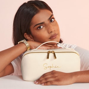 May include: Cream-colored makeup bag with a gold zipper and handle. The bag has the name "Sophie" in gold script. The bag is rectangular with rounded edges. The bag is sitting on a white surface.