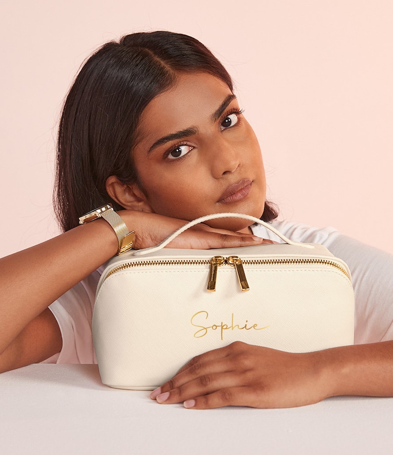 May include: Cream-colored makeup bag with a gold zipper and handle. The bag has the name "Sophie" in gold script. The bag is rectangular with rounded edges. The bag is sitting on a white surface.