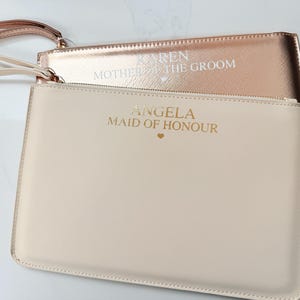 May include: Two rectangular wristlet pouches with wrist straps. The front pouch is cream-colored with gold text reading ANGELA MAID OF HONOUR and a small heart. The back pouch is metallic rose gold with white text reading KAREN MOTHER OF THE GROOM and a small heart.