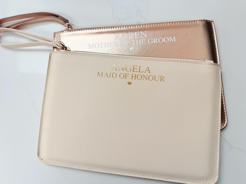 May include: Two rectangular wristlet pouches with wrist straps. The front pouch is cream-colored with gold text reading ANGELA MAID OF HONOUR and a small heart. The back pouch is metallic rose gold with white text reading KAREN MOTHER OF THE GROOM and a small heart.