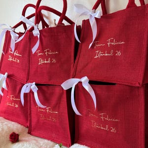 May include: Several red tote bags with matching handles and white ribbon bows. Each bag has the text "Team Felicia Istanbul 26" in gold script. The bags are stacked and arranged on a white surface.