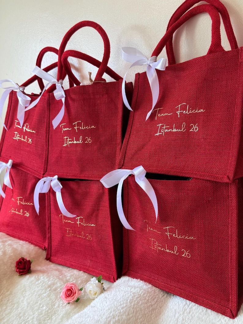 May include: Several red tote bags with matching handles and white ribbon bows. Each bag has the text "Team Felicia Istanbul 26" in gold script. The bags are stacked and arranged on a white surface.