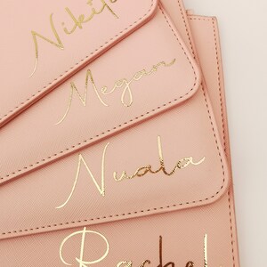 May include: Four pink pouches with gold lettering. The pouches are stacked on top of each other. The names Nikita, Megan, Nuala, and Rachel are written in gold script on the pouches.
