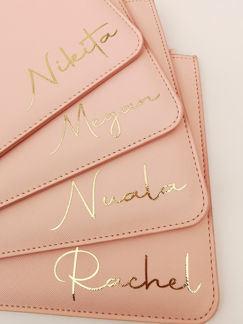 May include: Four pink pouches with gold lettering. The pouches are stacked on top of each other. The names Nikita, Megan, Nuala, and Rachel are written in gold script on the pouches.