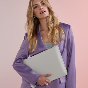 May include: A woman wearing a lavender blazer holds a light gray leather clutch with a gold zipper.