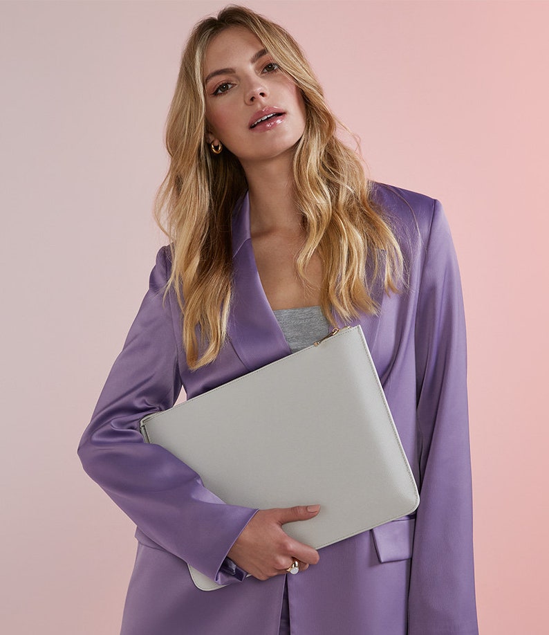 May include: A woman wearing a lavender blazer holds a light gray leather clutch with a gold zipper.