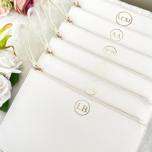 May include: Six white zippered pouches with gold handles and gold initials in a circle. The initials are: LB, LH, RL, KP, AA, and LCSJ.