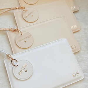 May include: A collection of four rectangular wristlet pouches in shades of white and beige. Each pouch features a gold-tone zipper, a matching wrist strap, a circular tag embossed with the text BRIDE or BRIDESMAID, and gold-stamped initials.