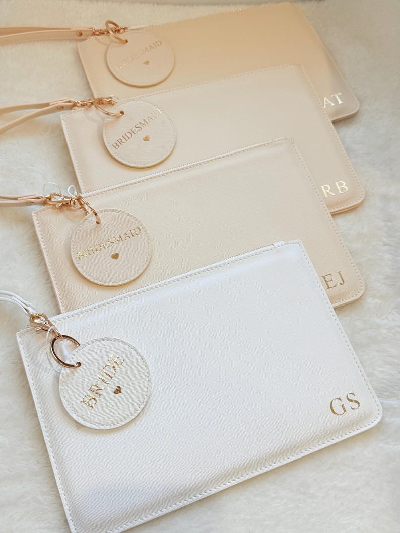 May include: A collection of four rectangular wristlet pouches in shades of white and beige. Each pouch features a gold-tone zipper, a matching wrist strap, a circular tag embossed with the text BRIDE or BRIDESMAID, and gold-stamped initials.