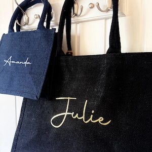 May include: Two tote bags, one navy blue and one black, hang on hooks. The navy bag has "Amanda" in white script, and the black bag has "Julie" in gold script. Both bags have sturdy handles.