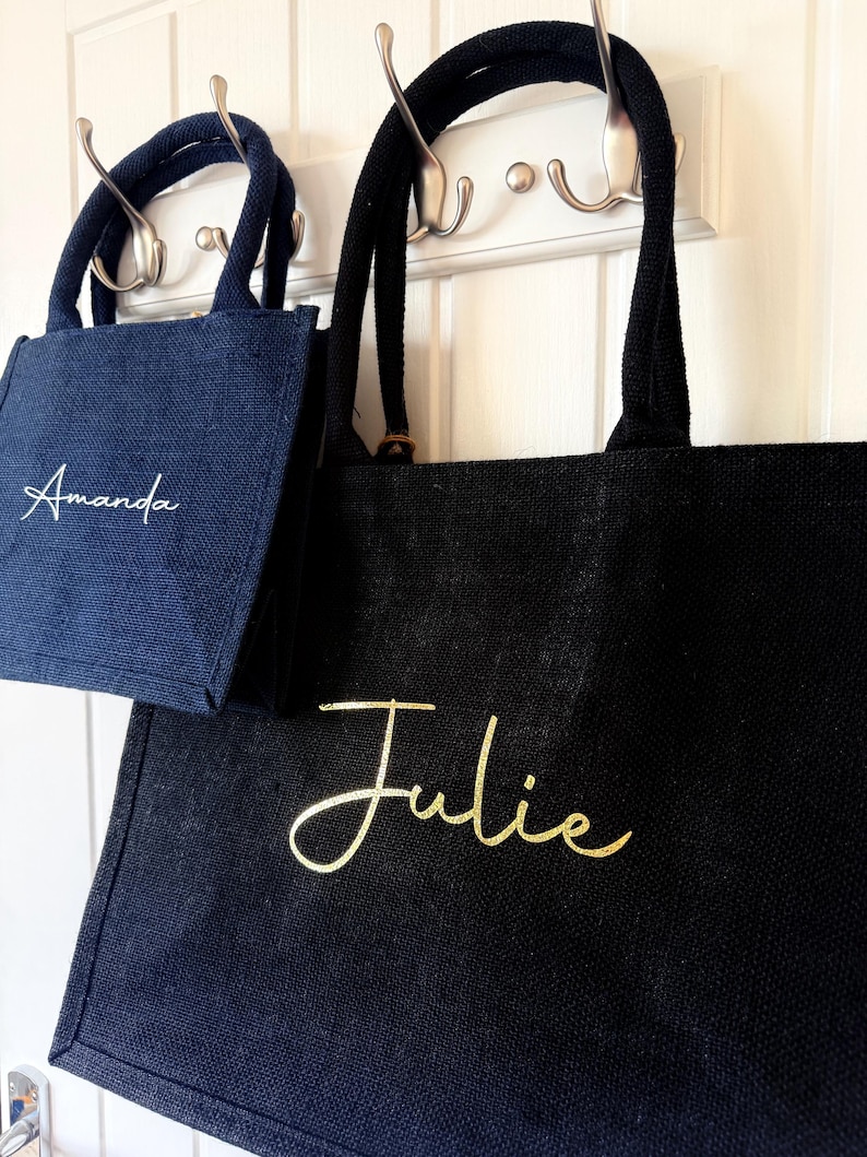 May include: Two tote bags, one navy blue and one black, hang on hooks. The navy bag has "Amanda" in white script, and the black bag has "Julie" in gold script. Both bags have sturdy handles.