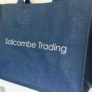 May include: A navy blue tote bag with the text "Salcombe Trading" in white. The bag has two handles and appears to be made of a textured fabric. The bag is rectangular and has a simple design.