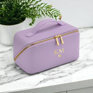 May include: A lavender makeup bag with a gold zipper and handle. The bag has the initials "KM" and a heart symbol in gold. The bag is sitting on a white marble surface, with a plant in the background.