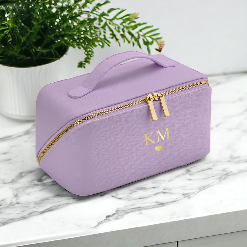 May include: A lavender makeup bag with a gold zipper and handle. The bag has the initials "KM" and a heart symbol in gold. The bag is sitting on a white marble surface, with a plant in the background.
