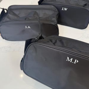 May include: Three black rectangular toiletry bags with zippered closures and top handles. Each bag features white embroidered initials on the front: J.A, J.W, and M.P. The bags are made of a smooth, durable fabric and have a structured, boxy shape.