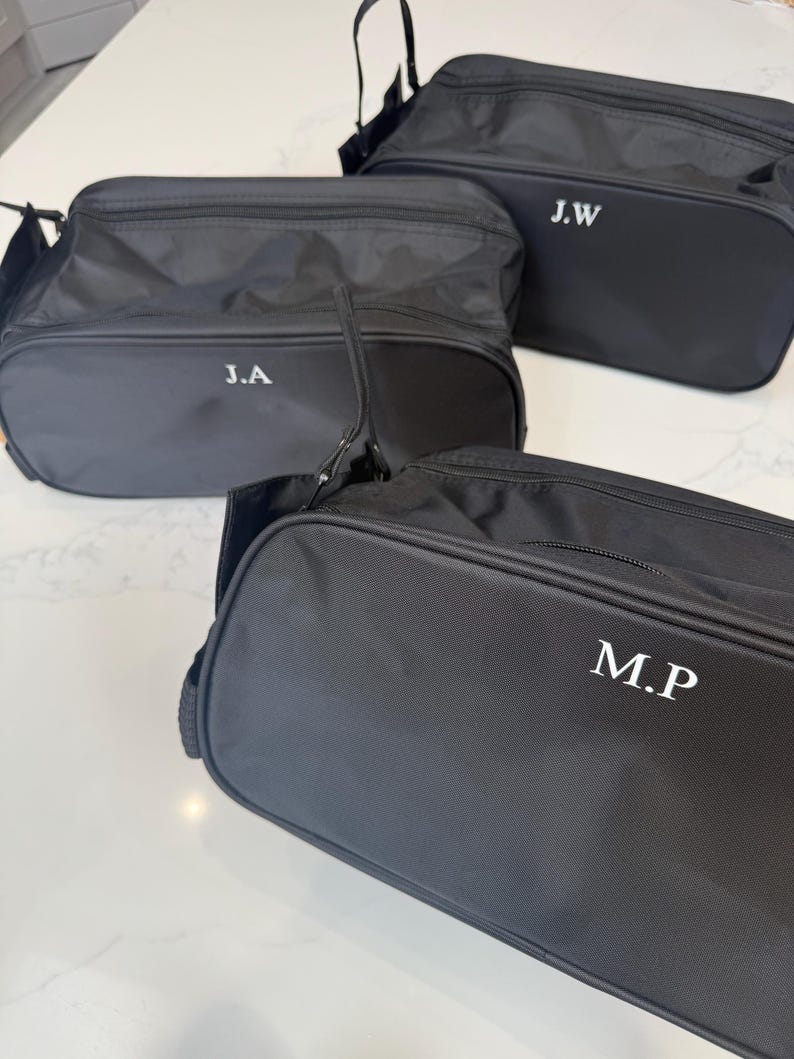 May include: Three black rectangular toiletry bags with zippered closures and top handles. Each bag features white embroidered initials on the front: J.A, J.W, and M.P. The bags are made of a smooth, durable fabric and have a structured, boxy shape.