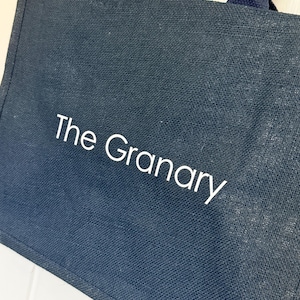 May include: Navy blue tote bag with the text "The Granary" in white. The bag has matching navy blue handles and a natural-colored jute string detail. The bag is made of a textured material.