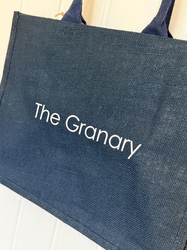 May include: Navy blue tote bag with the text "The Granary" in white. The bag has matching navy blue handles and a natural-colored jute string detail. The bag is made of a textured material.