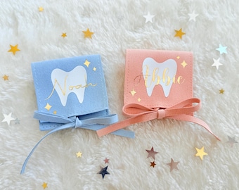 May include: Two small square pouches, one light blue and one peach, each featuring a white tooth graphic and gold cursive text reading Noah and Abbie. Each pouch is tied with a matching ribbon and decorated with small gold star accents.