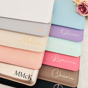 May include: A stack of ten zippered pouches in various colors, each with a different name or initials written in gold lettering. The pouches are made of a soft, textured material and have a subtle sheen.