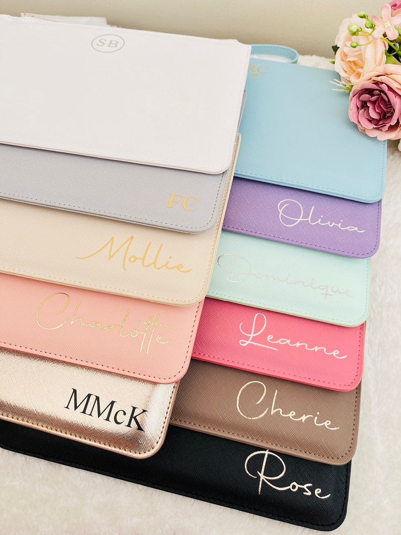 May include: A stack of ten zippered pouches in various colors, each with a different name or initials written in gold lettering. The pouches are made of a soft, textured material and have a subtle sheen.